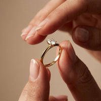 Gold ring with a diamond held between two fingers against a beige background Gold